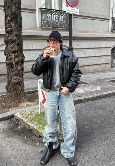 Una persona joven con gorro negro, gafas, chaqueta de cuero, suéter gris y vaqueros rotos y holgados, tomando café en la acera de la ciudad cerca de un árbol y una señal de tráfico.