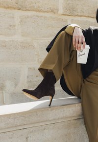 Personne portant un pantalon marron et des bottines à talons foncées assise sur un rebord en pierre, tenant une tasse de café blanche de Café Kitsuné.