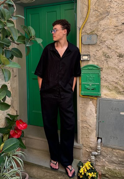 Camisa negra de manga corta y pantalones holgados, llevados con chanclas, de pie junto a una puerta verde y una pared de piedra texturizada, rodeado de plantas.