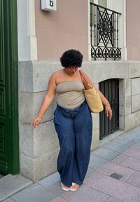 Smėlinės spalvos be petnešėlių viršus, tamsiai mėlynos laisvo silueto kelnės ir smėlinės spalvos rankinė. Moteris su trumpais garbanotais plaukais stovi ant šaligatvio prie žalios durų.