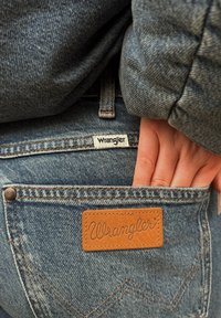 Modré džíny z denimu s koženou nášivkou označenou "Wrangler", s kontrastním šitím a rukou částečně zasunutou v zadní kapse.