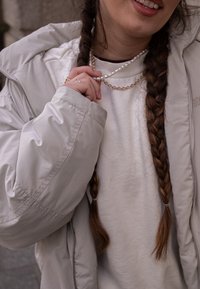 Casaco puffer bege com um colarinho alto, usado sobre um sweatshirt de cor clara. Colares em camadas de ouro e pérolas visíveis. Cabelo castanho trançado.
