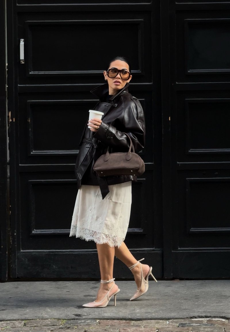 Woman wearing oversized black leather jacket, white lace skirt, beige heels, and large sunglasses, holding a coffee cup and brown handbag.