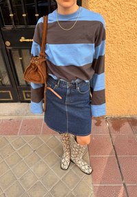 Personne portant un pull à rayures bleues et marron, une jupe en jean, des bottes à imprimé serpent, un collier en or et portant un sac bandoulière en daim marron.