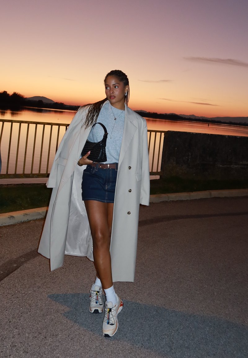 Ljusbeige kappa, grå topp, jeanskjol, vita strumpor, klumpiga sneakers med flerfärgade accenter och en liten svart handväska. Solnedgång i bakgrunden.