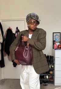 Άτομο που φοράει καρό καπέλο και μεγάλη σακάκι, κρατώντας μια μεγάλη μπορντό τσάντα, στέκεται σε ένα δωμάτιο με παλτό και παπούτσια στο παρασκήνιο.