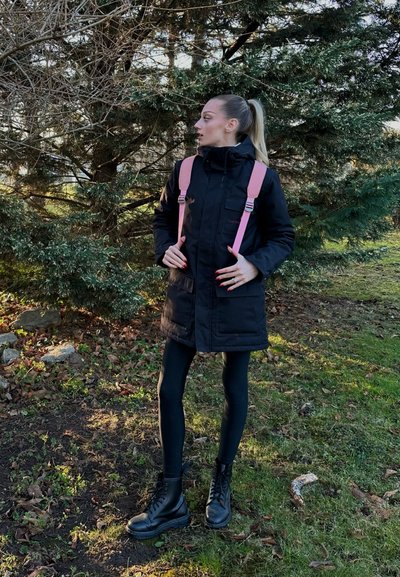 Abrigo acolchado negro con correas rosas, mangas largas ajustadas, bolsillos delanteros, leggings negros y botas negras gruesas, situado en un entorno exterior verde.