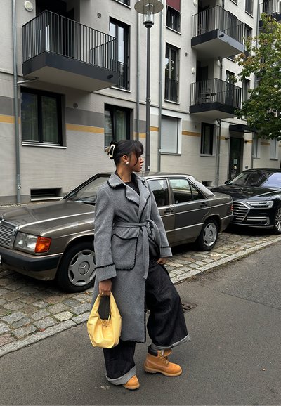 Abrigo de lana gris con acentos negros y cinturón, combinado con pantalones de pierna ancha oscuros. Bolso amarillo y botas beige. Entorno urbano con coches.
