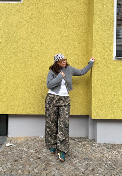 Joven con cabello rizado, gafas, suéter gris, pantalones con estampado y zapatillas, de pie frente a una pared texturizada de color amarillo brillante.