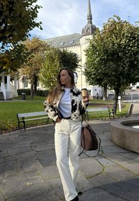 Kurtka z futerkiem ze sztucznego materiału w czarno-biały wzór, biała koszulka, białe jeansy z wysokim stanem, czarny pasek, brązowa torba, trzyma kubek kawy na zewnątrz.