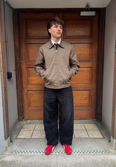 Chaqueta de aviador marrón, pantalones de pierna ancha negros y zapatillas rojas. Fondo de puerta de madera, texturas suaves y elementos de diseño simples.