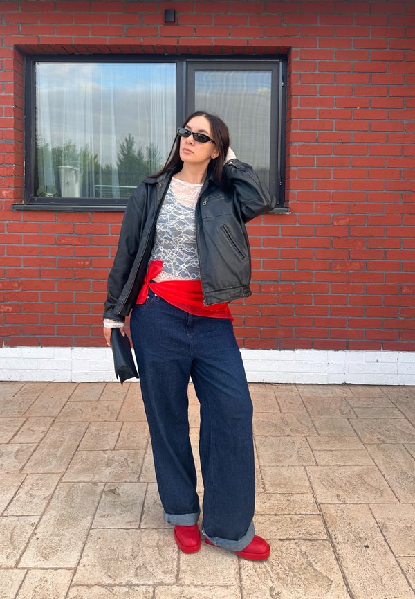 Sorte læderjakke, blonde top, rød bælte, brede blå jeans, røde sko; står foran en murstensvæg og vindue, hænderne i håret.