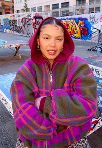 Giacca scozzese multicolore con tonalità viola, rosa e verde; cerniera anteriore; grandi maniche imbottite; indossata sopra una felpa con cappuccio coloramaranto.