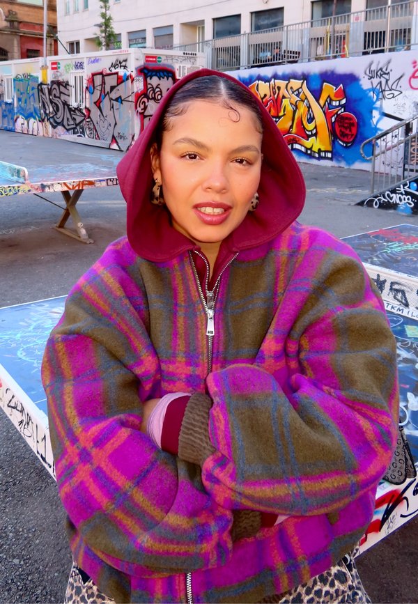 Chaqueta de cuadros multicolor con tonos morados, rosas y verdes; cierre frontal con cremallera; mangas grandes y abullonadas; llevada sobre una sudadera con capucha granate.