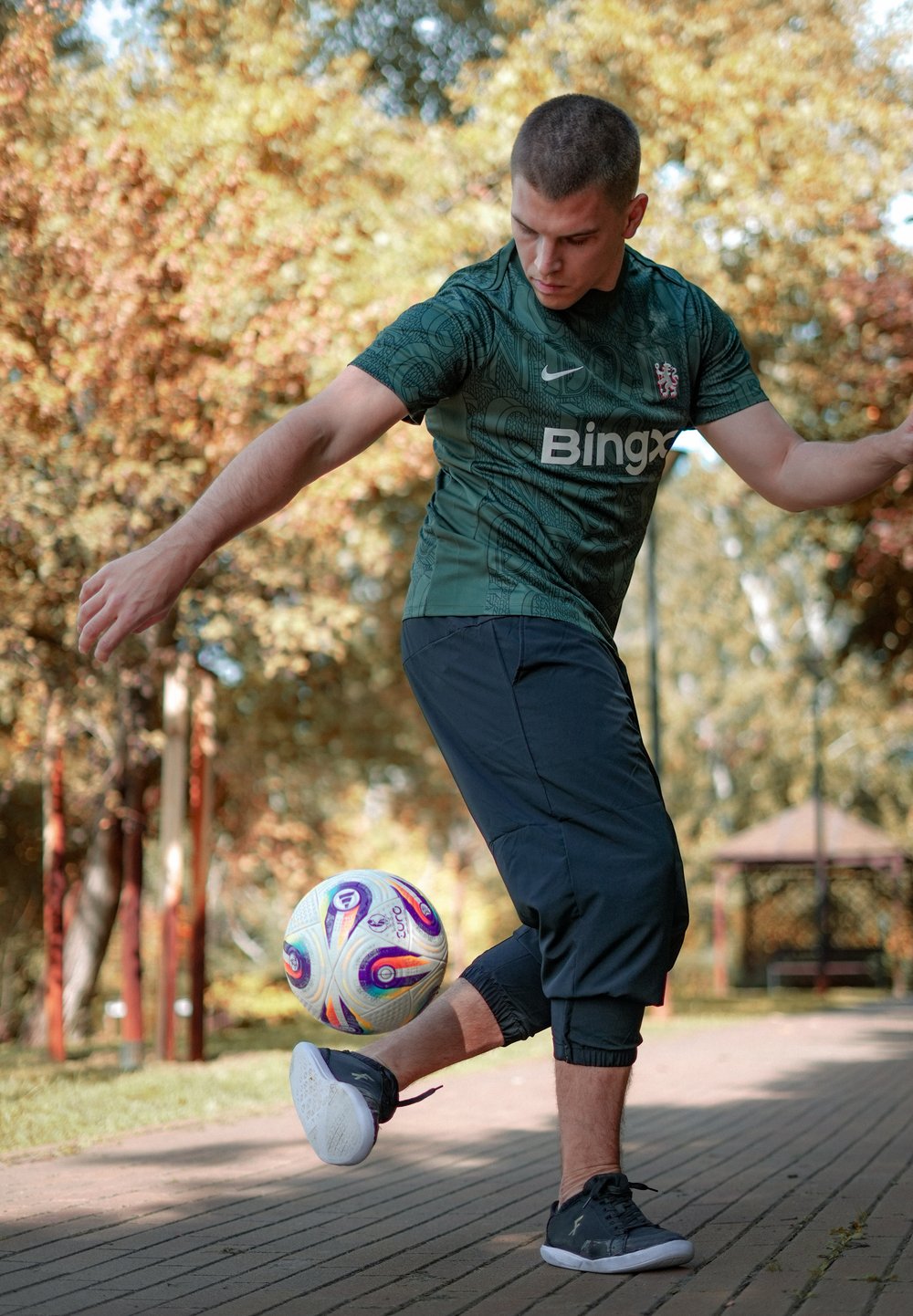 Zaļa sporta krekla ar teksturētu dizainu, melni sporta bikses un melnas čības. Izteiksmīgs ir krāsains futbols.