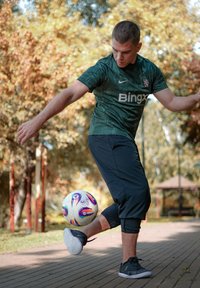 Zaļa sporta krekla ar teksturētu dizainu, melni sporta bikses un melnas čības. Izteiksmīgs ir krāsains futbols.