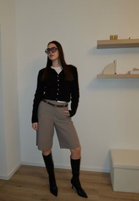 Jeune femme en cardigan noir, bermuda beige avec ceinture, bottes noires à talons montant jusqu'aux genoux et grandes lunettes, debout dans une pièce minimaliste.