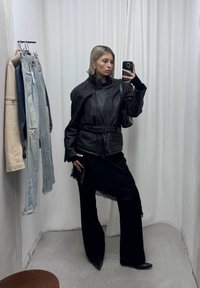 Personne aux cheveux blonds courts portant une veste en cuir noire oversize et un pantalon noir, prenant un selfie dans un miroir d'une cabine d'essayage.