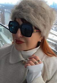 Femme aux cheveux roux portant de grandes lunettes de soleil noires, un chapeau en fourrure beige, un manteau beige, un col roulé blanc, des boucles d'oreilles en perles et des bagues près d'une rivière enneigée.