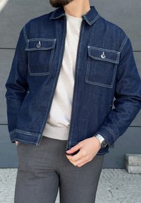 Chaqueta de mezclilla oscura con dos bolsillos en el pecho y costuras blancas en contraste; llevada sobre un suéter gris claro y pantalones grises; exhibida con luz natural.