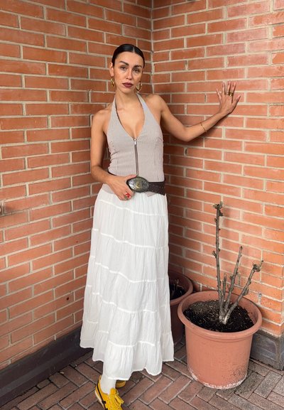 Top halter de rayas con cremallera, falda maxi blanca de volantes, cinturón marrón con hebilla y zapatillas amarillas. El fondo presenta una pared de ladrillo y una planta en maceta.