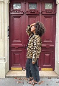 Jas met panterprint en zwarte afwerking, wijde zwarte broeken en bijpassende schoenen met panterprint. Bordeaurode deuren en stenen muur op de achtergrond.