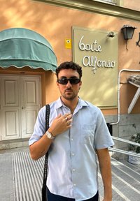 Camisa azul claro de mangas curtas com um bolso, óculos de sol pretos, relógio de pulso prateado e anéis. O fundo apresenta um toldo e uma placa.