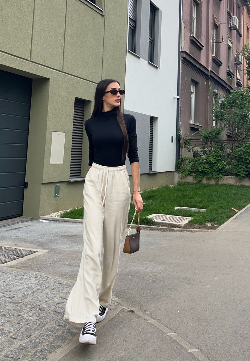 Vrouw loopt op straat met een zwart topje met lange mouwen, brede beige broek, zwarte sneakers, zonnebril en houdt een kleine bruine handtas vast.