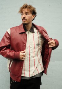 Burgundská umělá kožená bunda s bílými detaily, zapínání na knoflíky a žebrované manžety. Nese na sobě pruhované krémové a �červené tričko s kapsou na hrudi.
