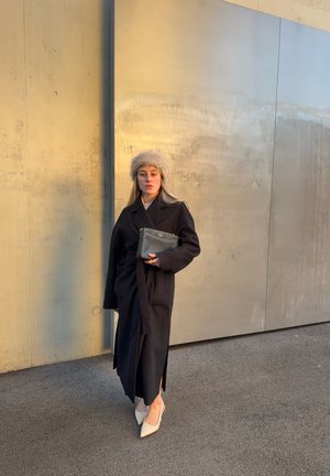 Mujer con abrigo largo negro, sombrero de piel beige, tacones blancos, sosteniendo un bolso de mano negro, de pie contra una pared beige texturizada al aire libre.