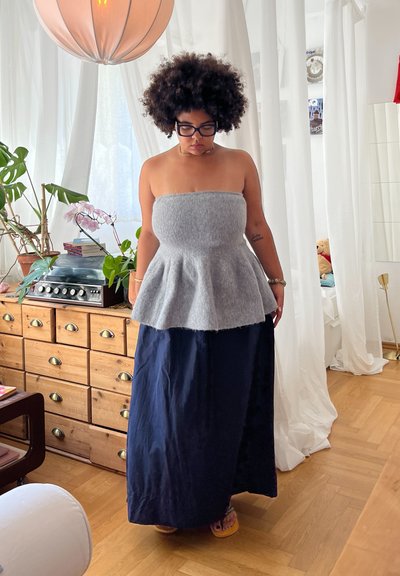 Top de peplum sin tirantes en color gris, hecho de material suave, combinado con una falda larga de color marino. Muebles de madera y plantas visibles en el fondo.