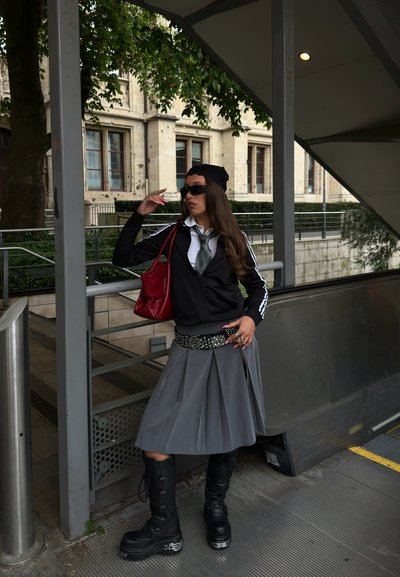 Joven mujer con chaqueta negra, falda gris y botas parada junto a una barandilla al aire libre, ajustándose las gafas de sol, llevando un bolso rojo bajo la escalera.
