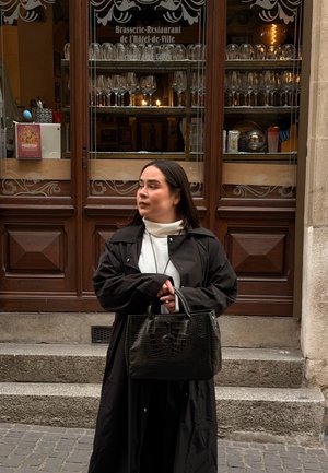 Mujer con abrigo negro y bolso negro está de pie en las escaleras fuera de una brasserie-restaurante con cristalería expuesta detrás de puertas de madera.