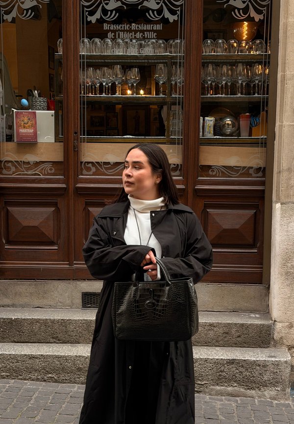 Kvinde i sort frakke med sort håndtaske står på trapperne udenfor en brasserie-restaurant med glasvarer udstillet bag trædøre.