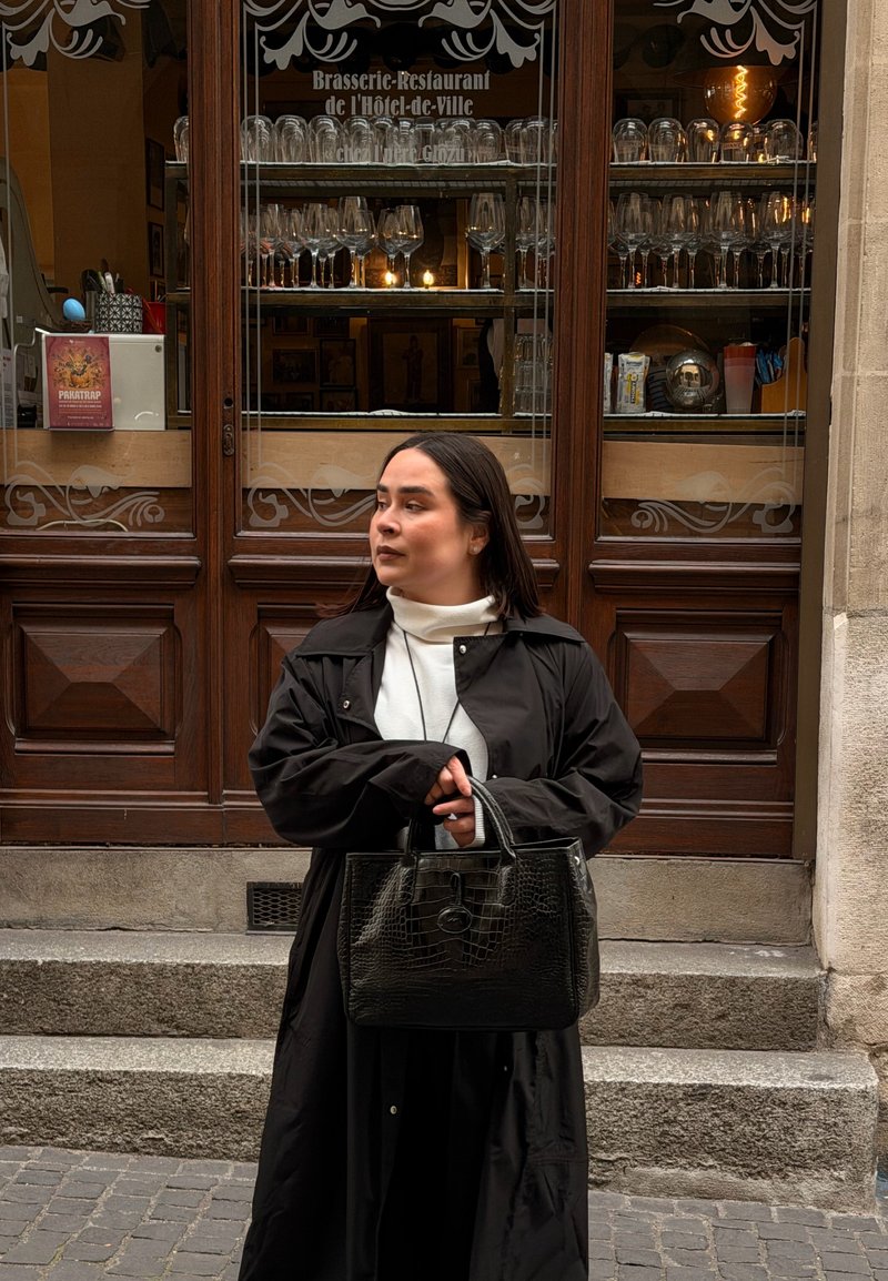 Femme en manteau noir tenant un sac à main noir, debout sur les marches devant un brasserie-restaurant avec de la verrerie exposée derrière des portes en bois.
