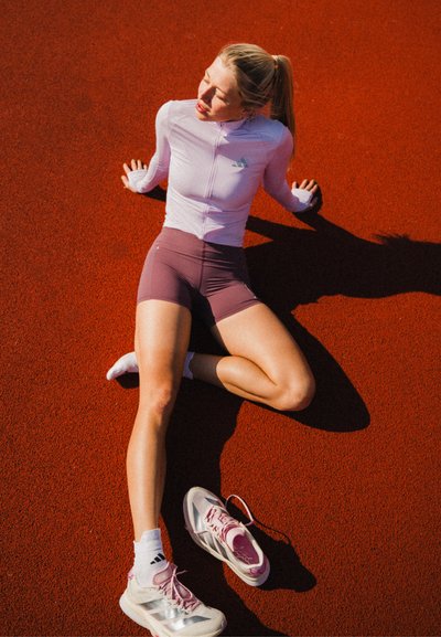 Mujer con ropa deportiva sentada en una pista roja, mirando hacia arriba con los ojos cerrados, con un zapato quitado a su lado.