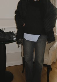 Châle oversize rayé noir avec bords à franges, superposé sur un pull foncé et une chemise bleu clair. Porté avec un jean évasé gris.