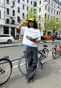 Personne portant une casquette néon et des lunettes de soleil, lisant un livre en s'appuyant sur un vélo dans une rue de la ville avec des bâtiments et des vélos stationnés.