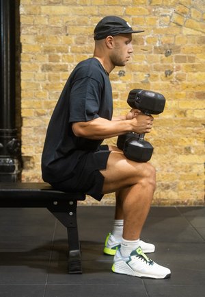 Black t-shirt and shorts worn by a person seated on a bench, holding a black dumbbell. The background features exposed brick walls.