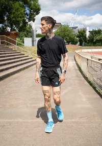Indivíduo atlético com uma camisa preta de mangas curtas e calções pretos com riscas verdes, usando ténis azuis e meias azuis, com tatuagens visíveis.