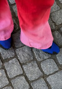 Kék cipők, textúrált felülettel és kivágott dizájnnal, rózsaszín ombre nadrággal párosítva, macskaköves járdán állva.