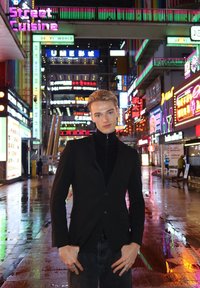 Giacca nera con zip e colletto alto, bottoni d'argento e jeans scuri. Lo sfondo mostra vivaci insegne al neon in una strada cittadina.