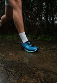 Jambes inférieures d'un coureur avec des chaussettes blanches et des chaussures de course Hoka bleues posant sur un sentier extérieur mouillé.