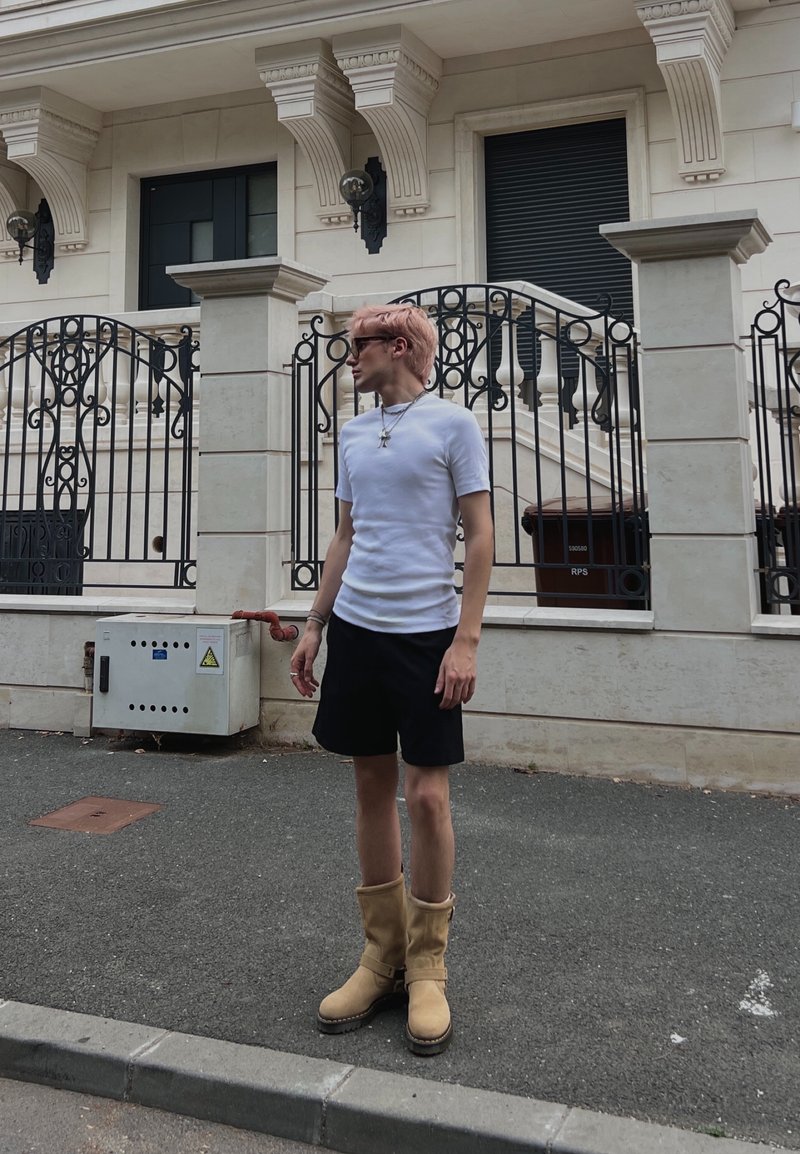 Jeune personne aux cheveux roses portant des lunettes de soleil, un t-shirt blanc, un short noir et des bottes couleur beige, debout dans une rue devant une clôture en fer décorative.