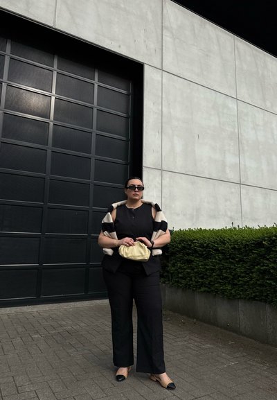 Conjunto negro que incluye una blusa abotonada y pantalones de pierna ancha, complementado con un chal a rayas. Sosteniendo un bolso de mano crema, de pie contra una pared gris.