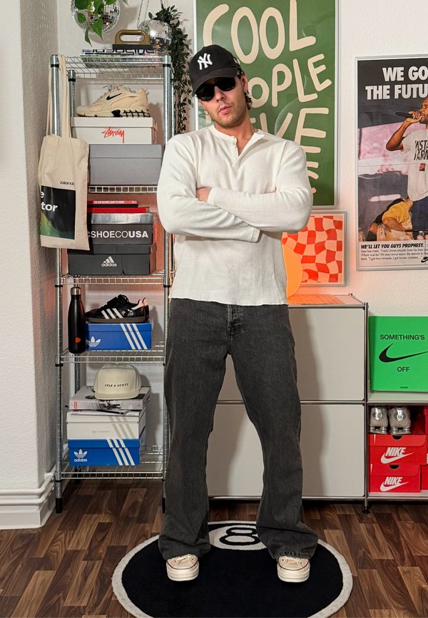 Un hombre con una gorra de los Yankees y gafas de sol está de pie con los brazos cruzados en una habitación llena de zapatillas deportivas. Lleva una camiseta blanca y vaqueros oscuros.