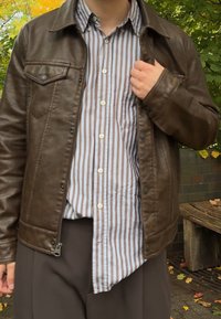 Asmuo vilki rudą odinę striukę ant dryžuotos marškinėlių, prisitaiko striukę lauke šalia medinio suolo ir rudeninių lapų.