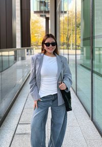 Blazer grigio sopra una camicia bianca, abbinato a jeans a gamba larga di un blu chiaro. Occhiali da sole neri e una borsa nera completano l'outfit.
