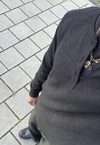 Pessoa vestindo uma camisa de botão de manga longa preta, colares de ouro, calças escuras e sapatos pretos, de pé sobre pavimentos de ladrilhos cinzentos.