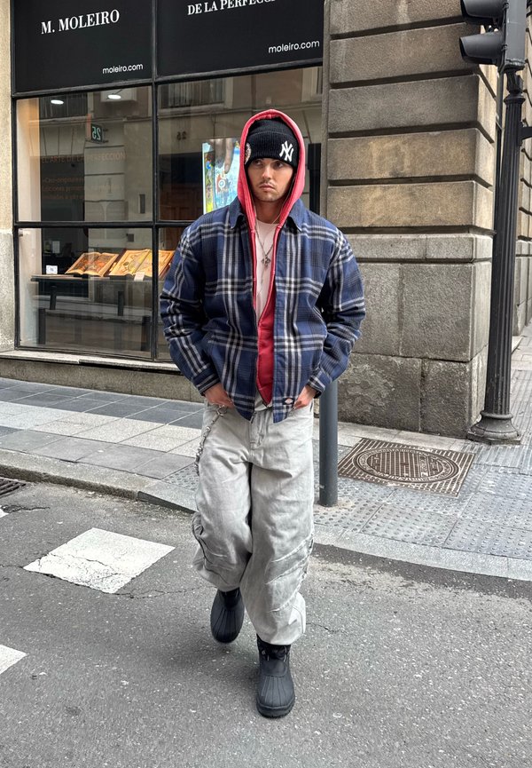 Bărbat tânăr care merge pe strada orașului purtând o cagulă neagră New York, hanorac roșu sub o jachetă albastră în carouri, pantaloni largi gri deschis și cizme negre.